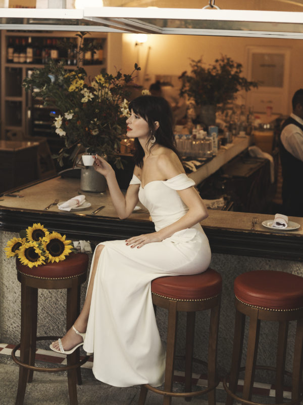 Bride sitting on stool in cafe wearing a draped off-the-should sheath wedding dress
